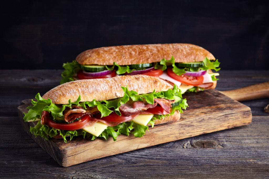 Two fresh submarine sandwiches with ham, cheese, bacon, tomatoes, lettuce, cucumbers and onions on wooden cutting board