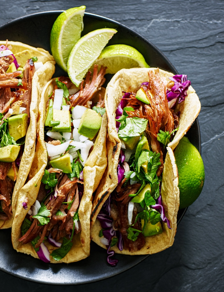 Pulled pork tacos with fresh veggies and cilantro, served on a dark plate with lime wedges.
