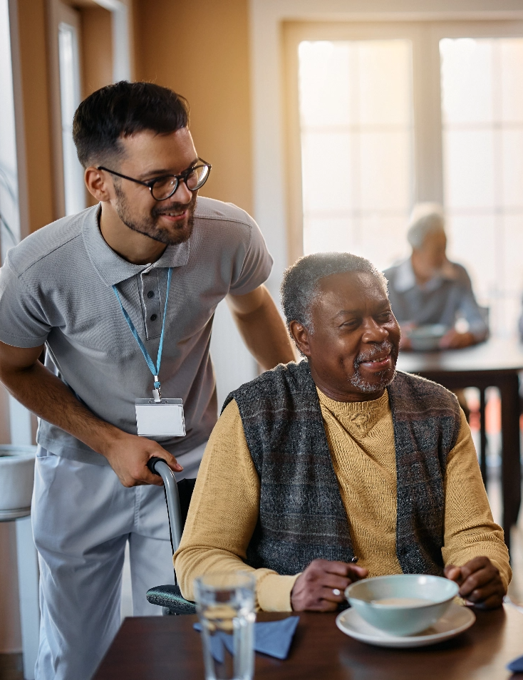 A senior living aide helps push a resident's wheelchair while both smile.