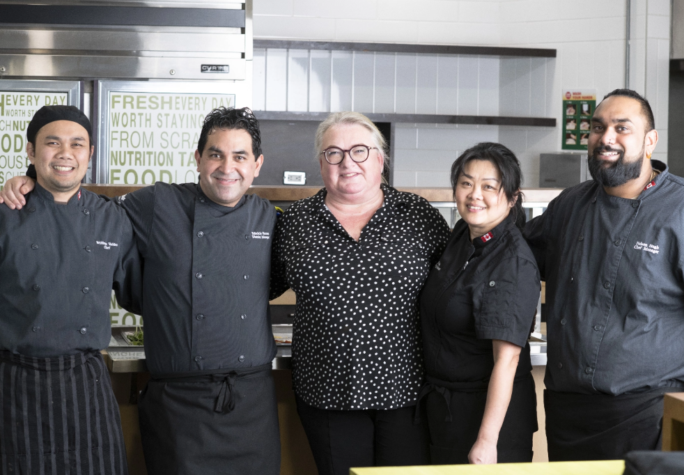 Un groupe d’employés de cuisine et de gestionnaires souriants pose devant une aire de préparation des aliments.