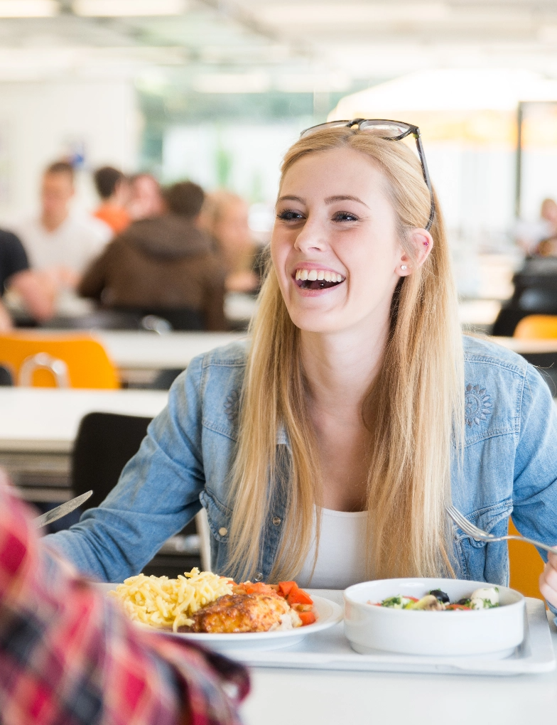 A young female student laughs with a classmate while eating fresh food off a tray in a cafeteria.