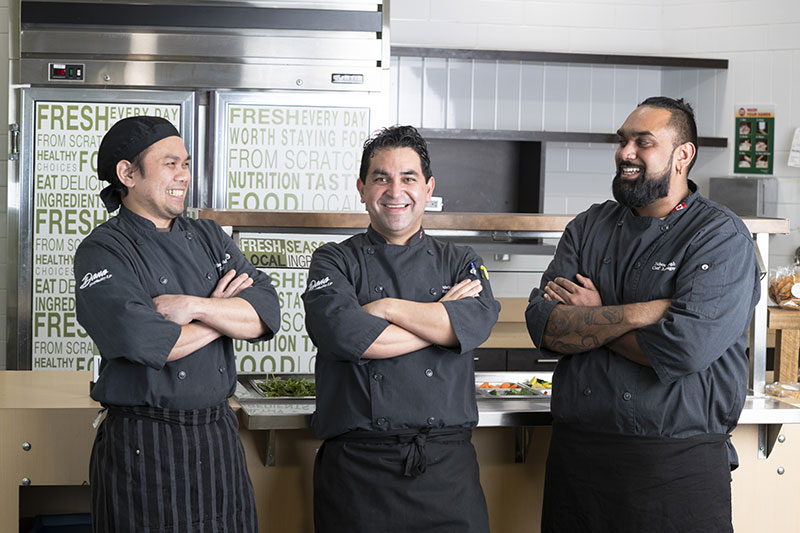 A group of smiling kitchen staff poses in front of a food preparation area.