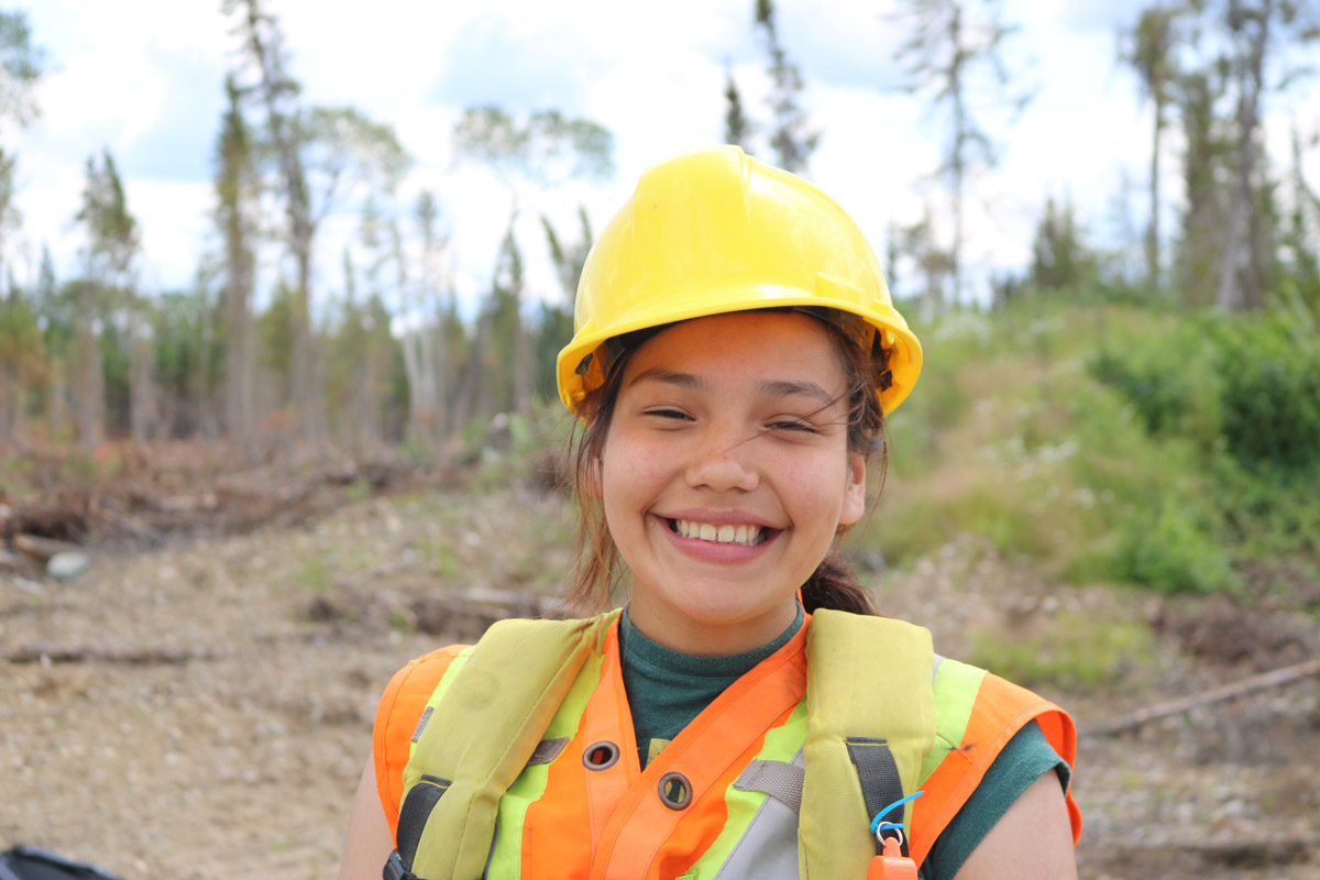 Une jeune femme qui porte un casque de protection et un gilet de sécurité et qui sourit à la caméra en se tenant debout sur un chantier de construction